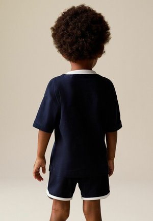 Göndör hajú gyermek áll háttal, tengerészkék ingben és fehér szegéllyel ellátott rövidnadrágban, egyszínű bézs háttér előtt.