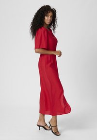 Robe rouge, à longueur genou, avec des manches courtes et bouffantes et une coupe décontractée. Tissu léger avec une texture lisse. Sandales noires à brides.