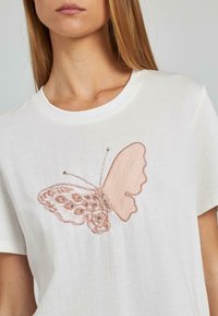 Camiseta blanca con un gran bordado de una mariposa rosa intrincada con adornos, escote redondo y mangas cortas.