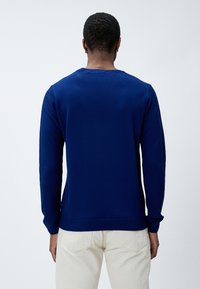 Granatowy sweter z dzianiny o rypsowym wzorze na rękawach i dolnym brzegu, okrągłym dekolcie i długich rękawach, zestawiony z jasnobrązowymi spodniami.