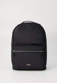 Mochila preta feita de tecido resistente, com formato arredondado, bolso frontal com fecho de correr e um detalhe metálico na frente. Textura suave.