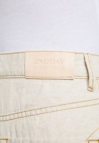 Beige denim pants feature a leather tag embossed with "2NDDAY THINKTWICE" and yellow stitching along the seams.
