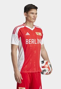 Maillot de football rouge et blanc à manches courtes, avec "BERLIN" en lettres majuscules. Tissu léger, motifs distincts et panneaux latéraux en maille.