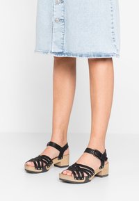 Zwarte sandalen met bandjes en houten blokhakken, voorzien van metalen studs, verstelbare enkelband en open teen ontwerp.