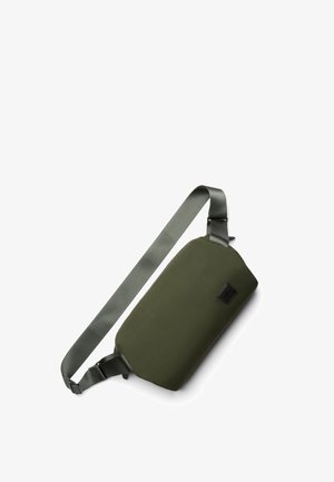 Sac bandoulière vert olive avec une finition lisse et mate, une sangle réglable et un petit patch logo noir sur le côté. Design minimaliste.