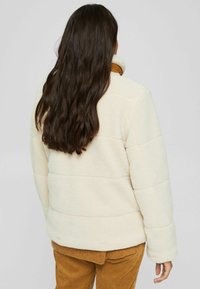 Kermas, fleece-takki korkean kauluksen ja tikatun kuvion kanssa. Takana on rento istuvuus, yhdistettynä ruskeisiin corduroy-housuihin.