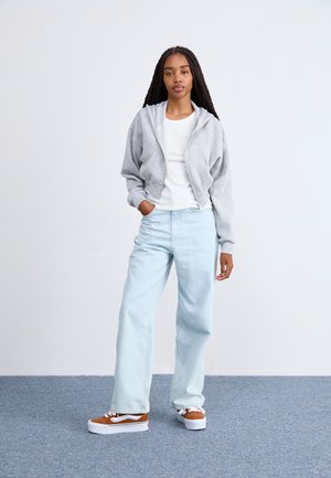 Felpa grigia con zip dal texture morbido, maglietta bianca a coste, jeans a gamba larga azzurri, e sneakers platform marroni con dettagli bianchi.