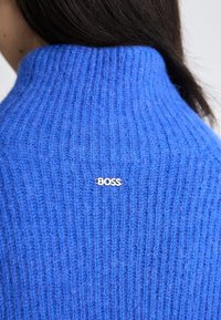 Blauer gerippter Strickpullover mit hohem Kragen und kleinem silbernen "BOSS"-Logoschild hinten in der Mitte. Weiche Textur und enganliegende Passform.