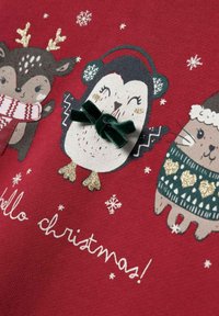 Un tessuto rosso presenta tre animali dei cartoni animati: un cervo marrone, un pinguino con un fiocco verde e un gatto in un maglione verde, con fiocchi di neve bianchi e testo.