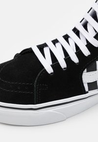 Czarne zamszowe sneakersy z białymi sznurówkami, z wzorem w szachownicę po bokach. Biała gumowa podeszwa z czarnymi akcentami.