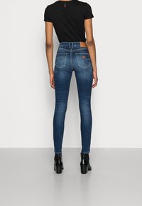 Vaqueros de mezclilla azul ajustados con una cintura alta, detalles de costura y una etiqueta de cuero marrón en la parte trasera de la cinturilla, combinados con botas de tacón negras.