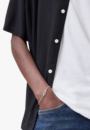 Camicia nera a maniche corte con bottoni color crema, indossata sopra una maglietta bianca a coste. Un braccialetto in argento adorna il polso, con jeans in denim blu visibili.