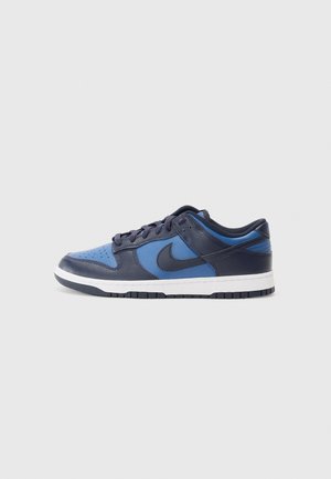 Lavprofil sneaker i blå og mørk marineblå med sort Nike-swoosh, hvid mellemsål og sort ydersål på en ensfarvet lys grå baggrund.