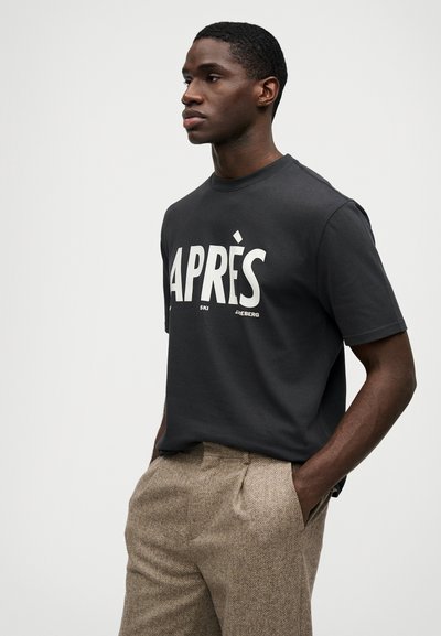 Σκούρο γκρι t-shirt με κοντό μανίκι που έχει λευκό κείμενο που λέει "APRÈS". Το μοντέλο το συνδυάζει με ανοιχτό καφέ καπιτονέ παντελόνι.