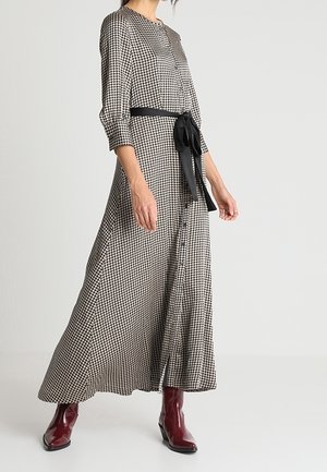Vrouw draagt een lange zwart-witte houndstooth-jurk met driekwartsmouwen, een zwarte tailleband en glanzende donkerrode enkellaarzen.