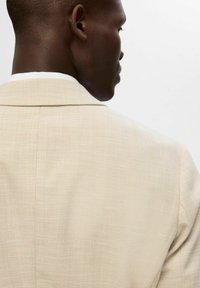 Beżowy blazer o teksturze z subtelnym wzorem w rybie łuski, z wykładanym kołnierzem i pojedynczymi szwami, widoczny z tyłu.