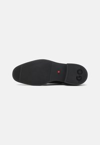 Semelle de chaussure en caoutchouc noir avec un motif de traction texturé, dotée d'un petit accent carré rouge et de bords arrondis. Comprend un logo en relief.