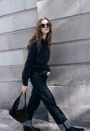 Sudadera con capucha oscura con cremallera, combinada con jeans de mezclilla de cintura alta con dobladillos enrollados, botines de cuero negro y un bolso marrón.