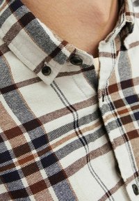 Geruite shirt met een beige, bruine en donkerblauwe kleurenschema. Zachte stof met zichtbare stiksels en twee zwarte knopen bij de kraag.