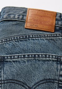 Jean Levi's 501 en denim bleu avec une surface texturée, présentant une patch en cuir marron au niveau de la taille avec des détails en relief.