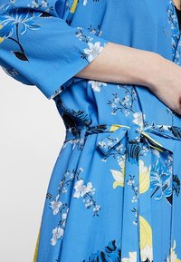 Modré šaty s květinovým vzorem a páskem v pase, s širokými rukávy a texturovanou látkou. V designu jsou výrazné žluté a černé barvy.