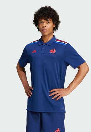 Polo-shirt bleu à manches courtes, avec des rayures rouges et bleues sur les épaules, un emblème de coq et le logo Adidas sur la poitrine.