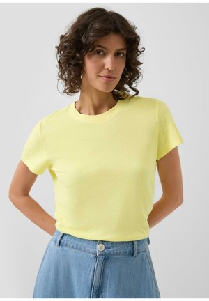 Femme aux cheveux bruns bouclés portant un t-shirt jaune à manches courtes et un jean bleu clair taille haute, mains derrière le dos, fond neutre.
