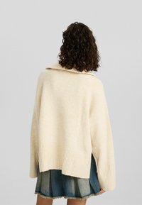 Cremefarbener gestrickter Pullover mit einem gerippten Saum und weitem Kragen, von hinten gezeigt, kombiniert mit einem gestuften blauen Jeansrock.