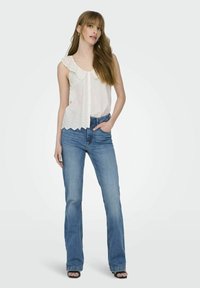 Blusa blanca sin mangas con borde de encaje en forma de onda y detalles de ojales, combinada con jeans de campana en azul claro y tacones negros destalonados.