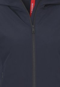 Giacca blu navy con una texture morbida, chiusura con zip frontale e un'etichetta di accento rossa al collo. Presenta un cappuccio e un design elegante.