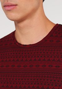 Röd t-shirt med ett geometriskt svart mönster, rund halsringning och korta ärmar. Tyget verkar mjukt med en texturerad tryckdesign.