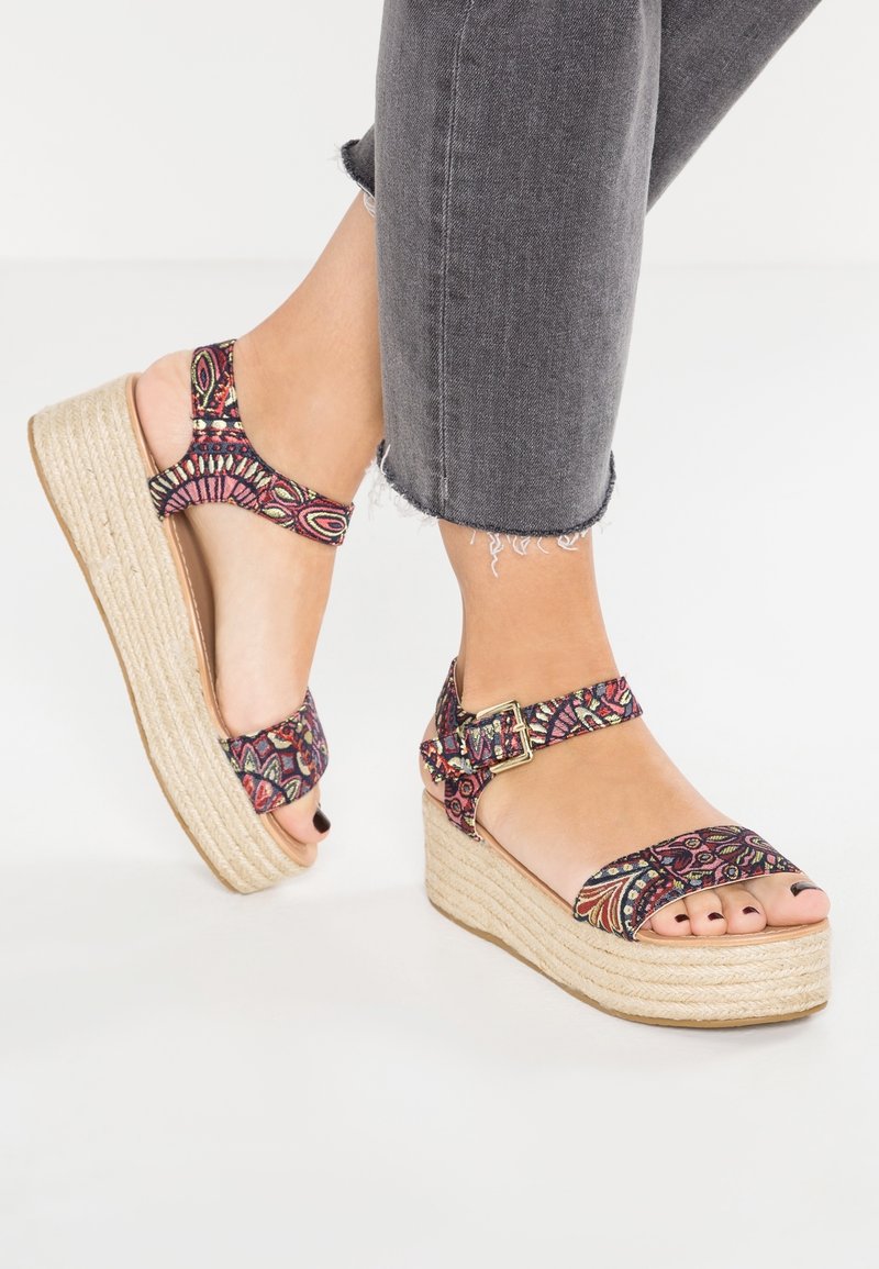 Pieds portant des sandales compensées avec des brides colorées à motifs et des boucles à la cheville, assorties à un jean noir raccourci.