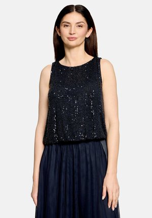 Femme aux longs cheveux foncés portant un haut noir sans manches à sequins et une jupe plissée bleu marine, debout devant un fond clair uni.