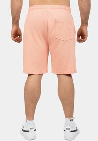 Shorts sportivi color pesca con elastico in vita, tasca posteriore e vestibilità comoda, abbinati a scarpe da ginnastica bianche con accenti neri.