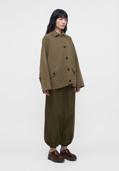 Mujer con chaqueta oversize de color verde oliva con botones, pantalones sueltos con puños y zapatos marrones de plataforma sin cordones, de pie frente a un fondo liso.