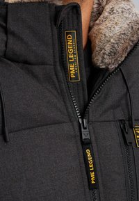 Čierna puffová bunda s golierom z umelého kožušiny, obsahujúca žlté značkové štítky, dvojité zipsy a textúrovanú tkaninu. Detailné prvky zahŕňajú čierne kovania.