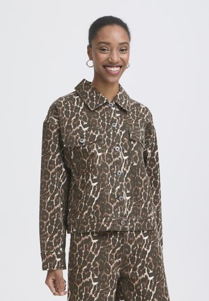 Kvinde, der smiler, iført matchende brun og sort leopardprintjakke og bukser, øreringe og ring på venstre hånd foran hvid baggrund.