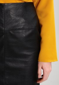 Ruka odpočívající po boku, oblečená v hořčicově žlutém rukávu a černé kožené sukni s viditelnými švy.