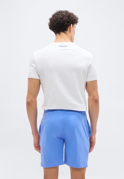 Άνδρας με την πλάτη προς τα εμάς φοράει λευκή κοντομάνικη μπλούζα με λογότυπο κοντά στη λαιμόκοψη και φωτεινά μπλε σορτς με τσέπες, στέκεται μπροστά σε απλό ανοιχτόχρωμο φόντο.