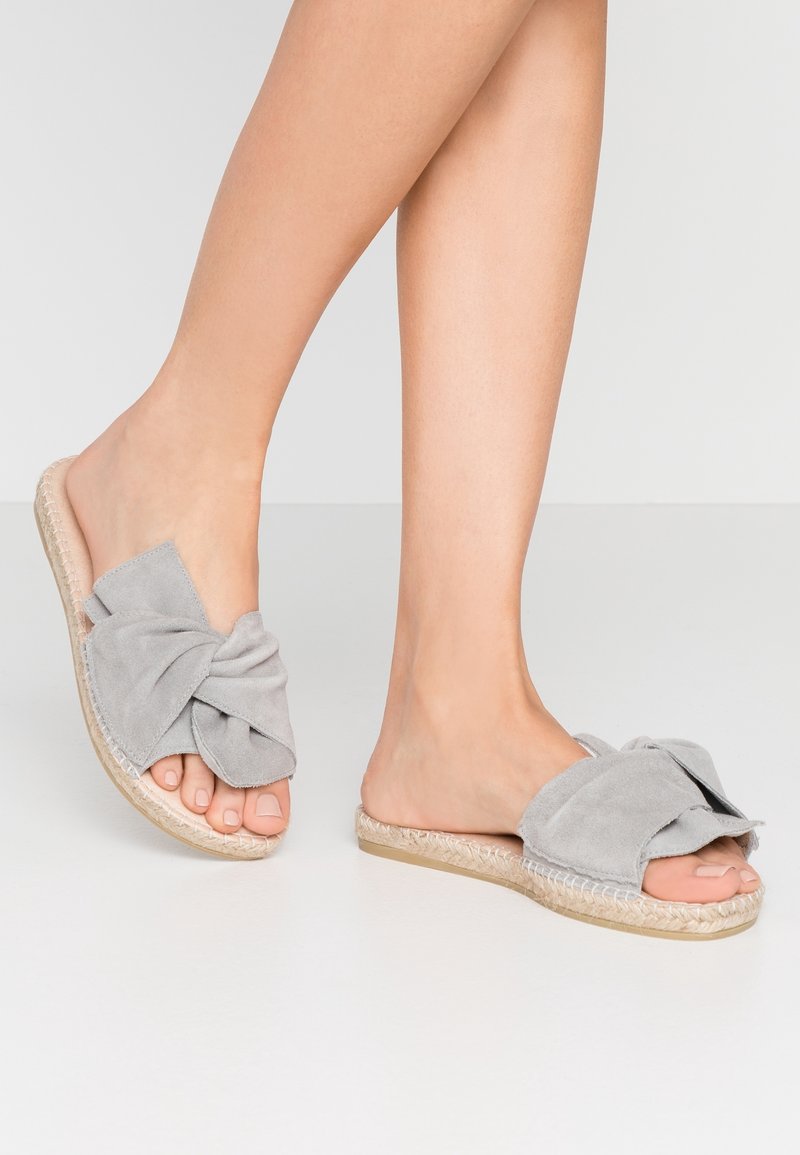 Pieds portant des sandales ouvertes à enfiler en daim gris clair avec de grands nœuds noués et des semelles en espadrilles tissées sur un sol blanc.