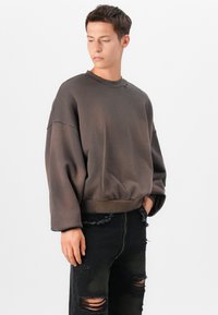 Sweatshirt oversized castanho com decote redondo e ombros caídos, combinado com calças jeans pretas desgastadas com rasgos.