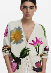 Cardigan en tricot avec une base crème et de grands imprimés floraux en rose, jaune et vert. Fermeture à cinq boutons à l'avant et poignets côtelés.