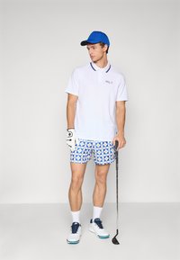 Hvit polo skjorte med blå-gul kant, blå mønstrede shorts, hvite golfsko, blå caps, og hvit hanske som holder en golfkølle.