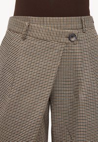 Primer plano de pantalones a cuadros marrones y beige con bolsillo lateral y cinturilla marrón con botones, combinados con una prenda superior oscura.