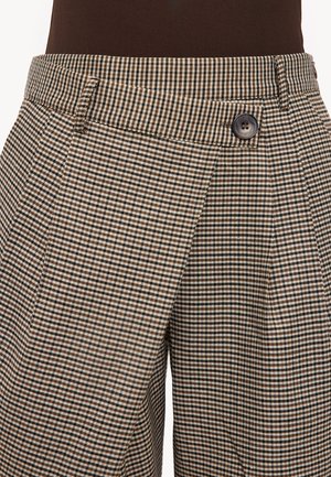 Close-up van bruin en beige geruite broek met zijzak en bruin met knopen sluitende tailleband, gedragen met een donker shirt.