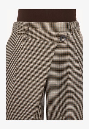 Gros plan sur un pantalon à carreaux marron et beige avec une poche latérale et une taille boutonnée marron, porté avec un haut foncé.
