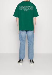 Grön oversized t-shirt med vit text "WRSTBHR STUDIO" på ryggen, kombinerad med ljusblå raka jeans och svarta mönstrade skor.