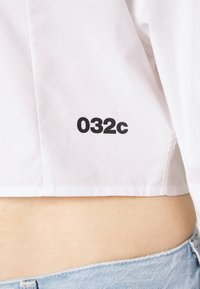 Witte cropped shirt van glad katoen, voorzien van een gedurfde zwarte "032c" print op de zijkant en een strakke, minimalistische uitstraling.