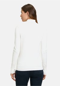 Weißer Langarm-Turtleneck-Pullover mit enganliegendem Design, gerippter Textur und hohem Kragen. Kombiniert mit dunklen, enganliegenden Hosen.