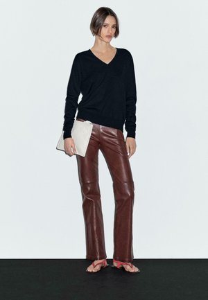 Das Model trägt einen schwarzen Pullover und braune Lederhosen, hält eine beige Clutch und steht vor einem schlichten Hintergrund mit einem ernsten Gesichtsausdruck.